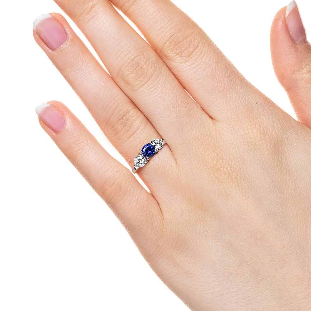 Shown with Round Cut Lab Grown Diamonds on either side of a Lab Created Blue Sapphire in 14k White Gold|Three stone engagement with round cut lab grown diamonds and lab created blue sapphire center stone in 14k white gold worn on hand