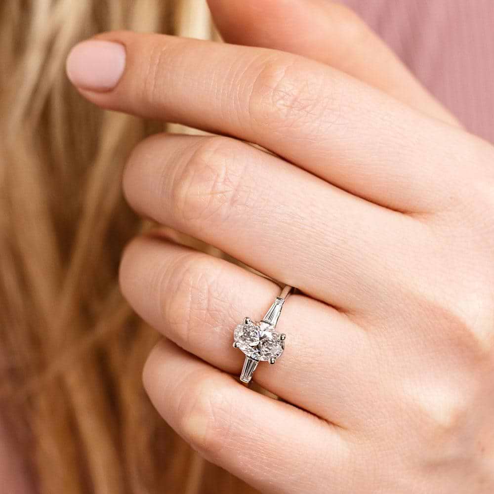 Shown with 2ct Oval Cut Lab Grown Diamond set in 14k White Gold|Beautiful three stone engagement ring with trellis set 2ct oval cut lab grown diamond amid baguette side stones in 14k white gold shown worn on hand