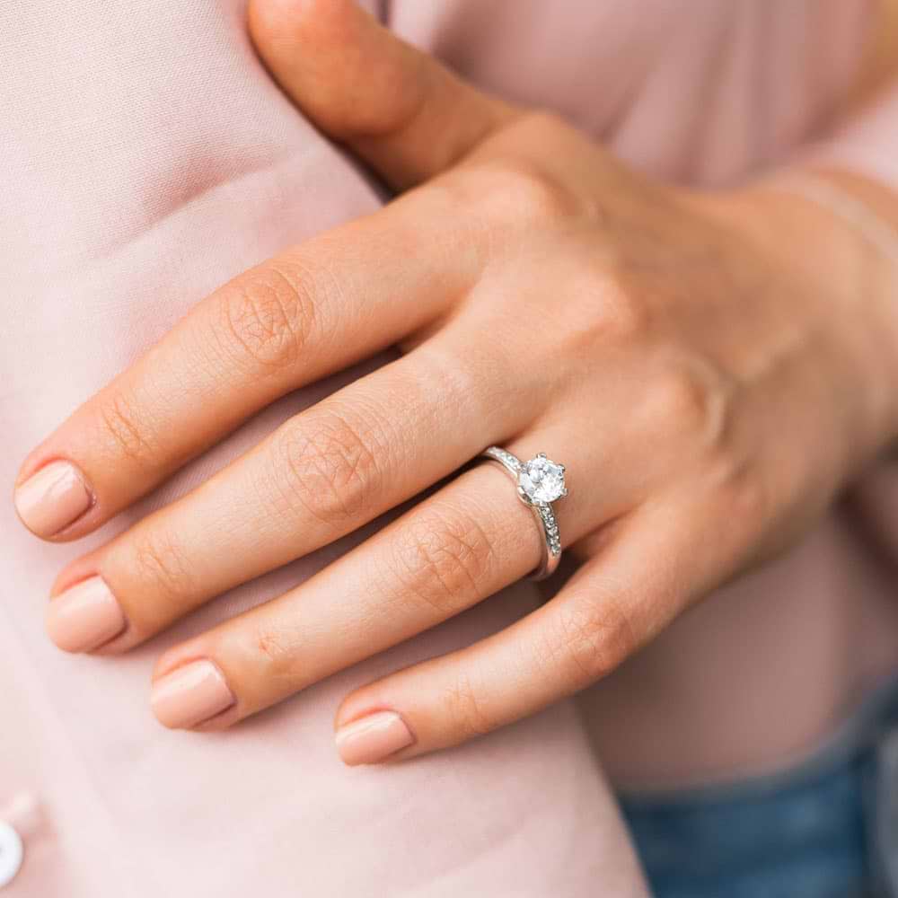 Shown with 1ct Round Cut Lab Grown Diamond in 14k White Gold|Channel set diamond accented engagement ring with 1ct round cut lab grown diamond in recycled 14k white gold worn on hand