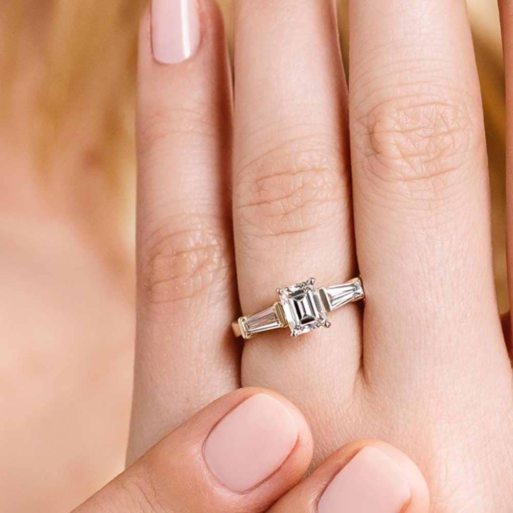 Shown with 1ct Emerald Cut Lab Grown Diamond in 14k Yellow Gold|Modern three stone engagement ring with two baguette cut recycled diamond side stones and a 1ct emerald cut lab grown diamond center in 14k yellow gold worn on hand