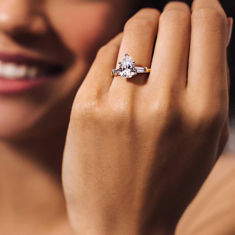 Shown with 2ct Pear Cut Lab Grown Diamond in 14k Yellow Gold|Beautiful three stone tear drop engagement ring with 2ct center stone in 14k yellow gold worn on hand