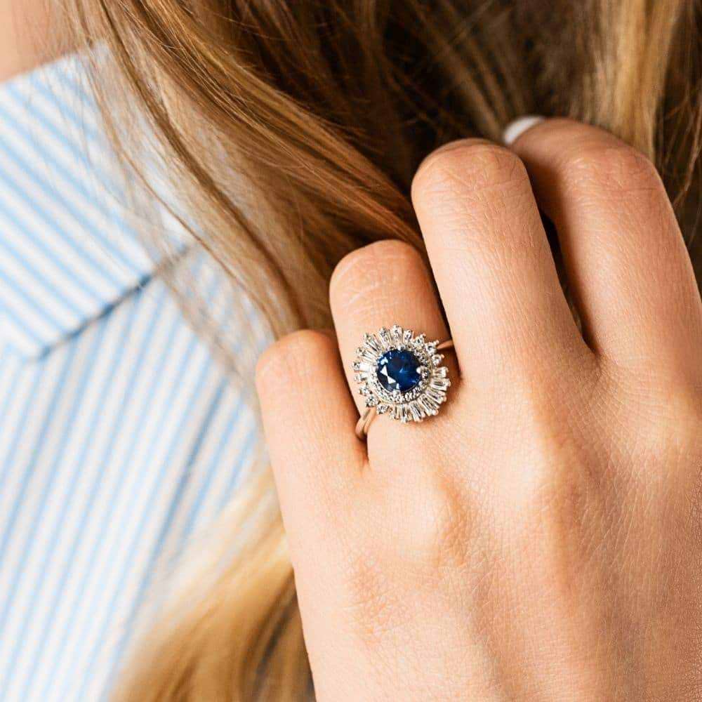 Shown with 1ct Round Cut Lab Grown Blue Sapphire|Unique stunning vintage style diamond halo engagement ring with baguette cut diamonds surrounding a 1ct round cut lab grown blue sapphire in 14k white gold worn on hand