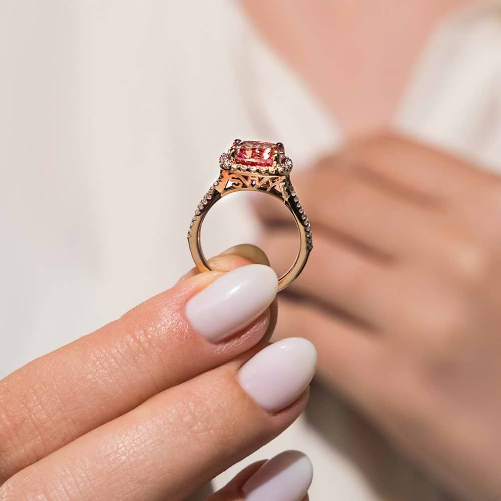 Shown with 1ct Cushion Cut Lab Grown Pink Sapphire in 14k Rose Gold|Halo monogram engagement ring with crossover split shank band set with 1ct cushion cut lab created pink sapphire in 14k rose gold shown held in hand