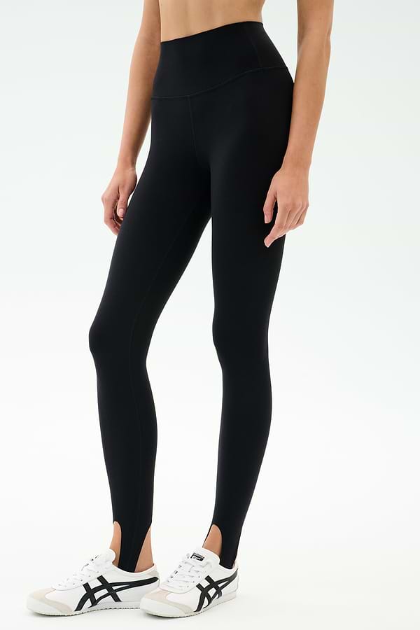 A person wears the SPLITS59 River High Waist Airweight Stirrup legging in black with white athletic shoes, standing against a plain light background.