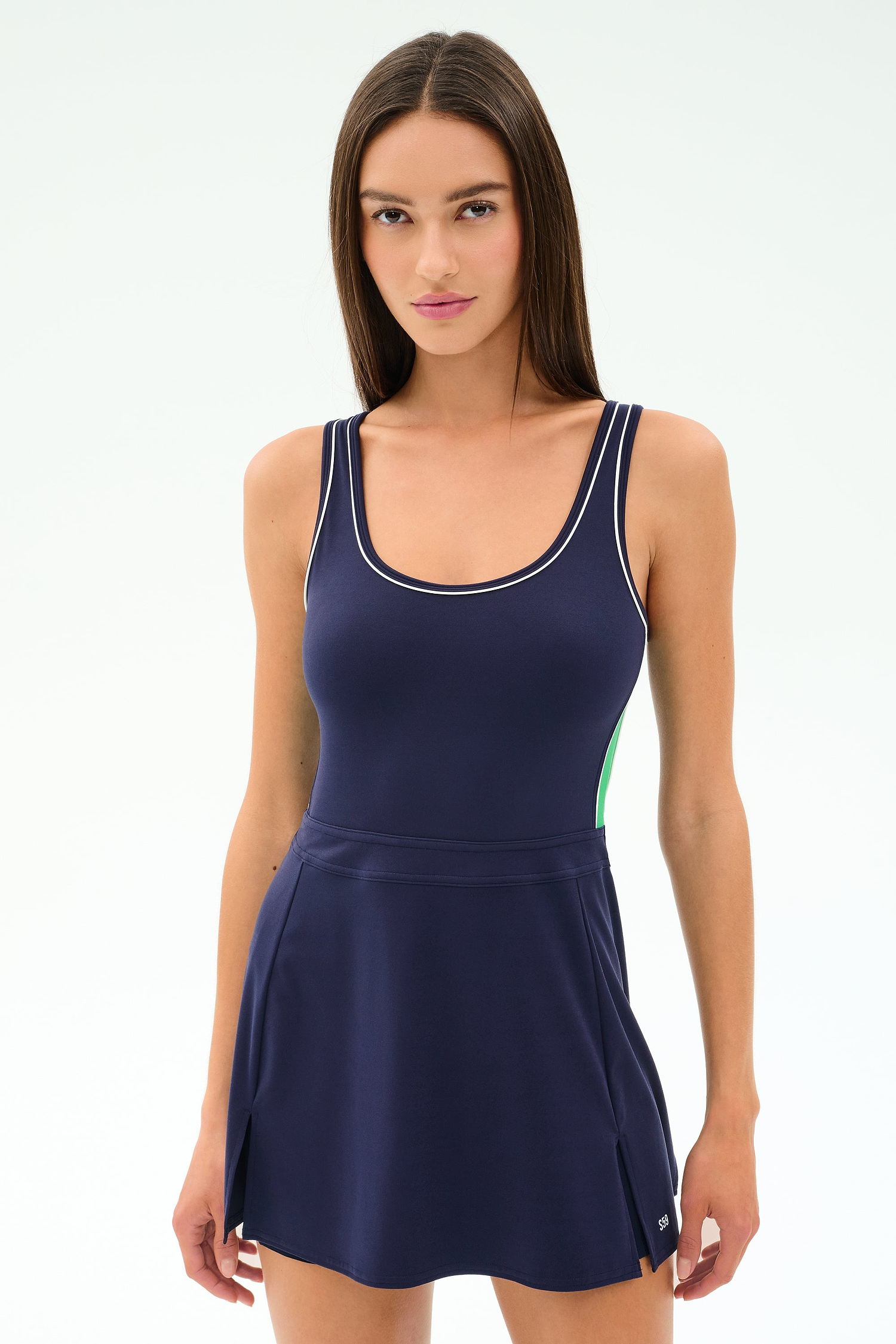A woman with long brown hair wears the SPLITS59 Martina Rigor Color Block Dress in Indigo/Spearmint, a navy sleeveless athletic style with white trim, made from a stretchy nylon-spandex blend, standing against a plain light background.