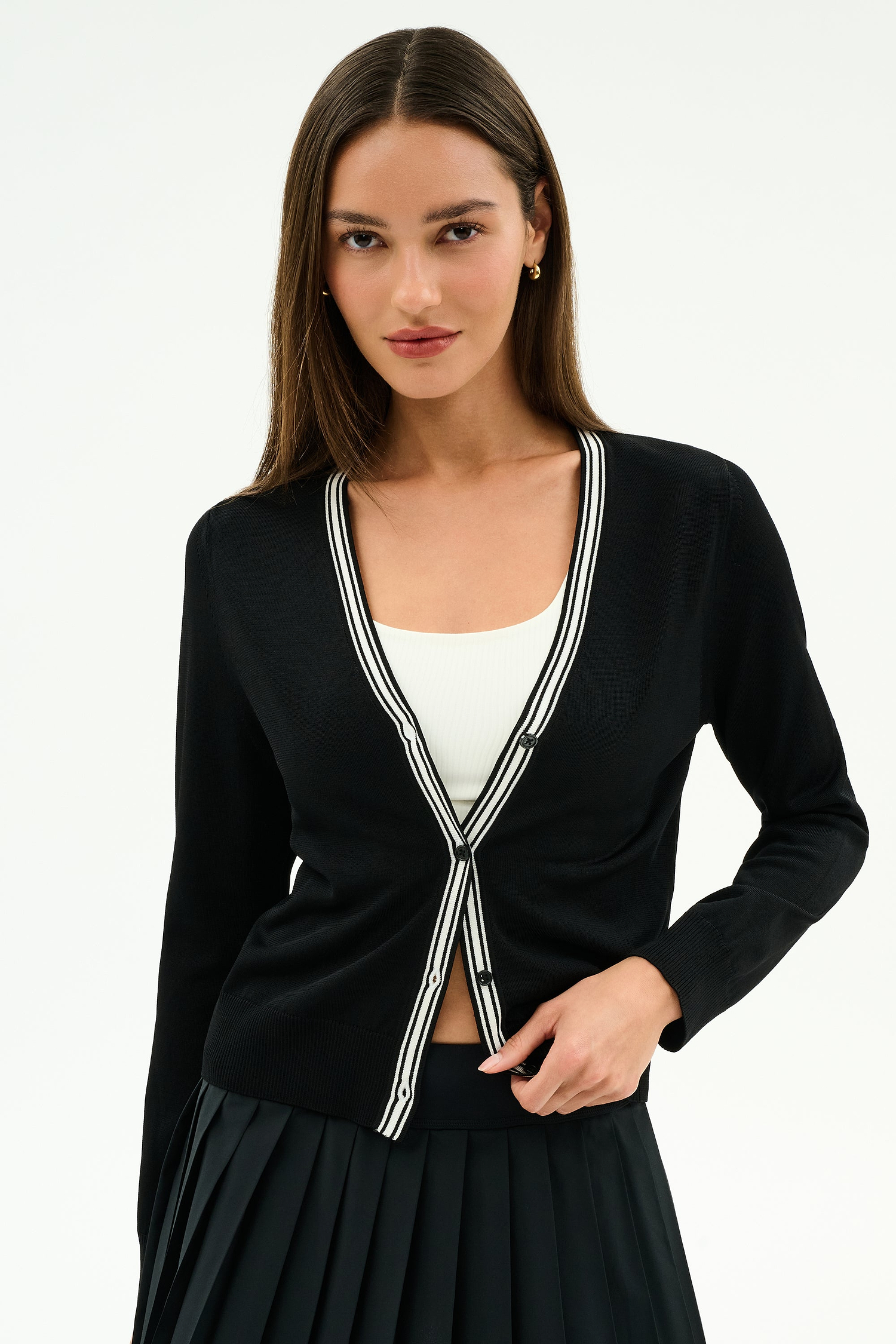 A woman models the SPLITS59 Bekah Knit Cardigan in Black/White—a soft rayon-nylon blend with white striped trim—over a white top and black pleated skirt, standing against a plain white background.