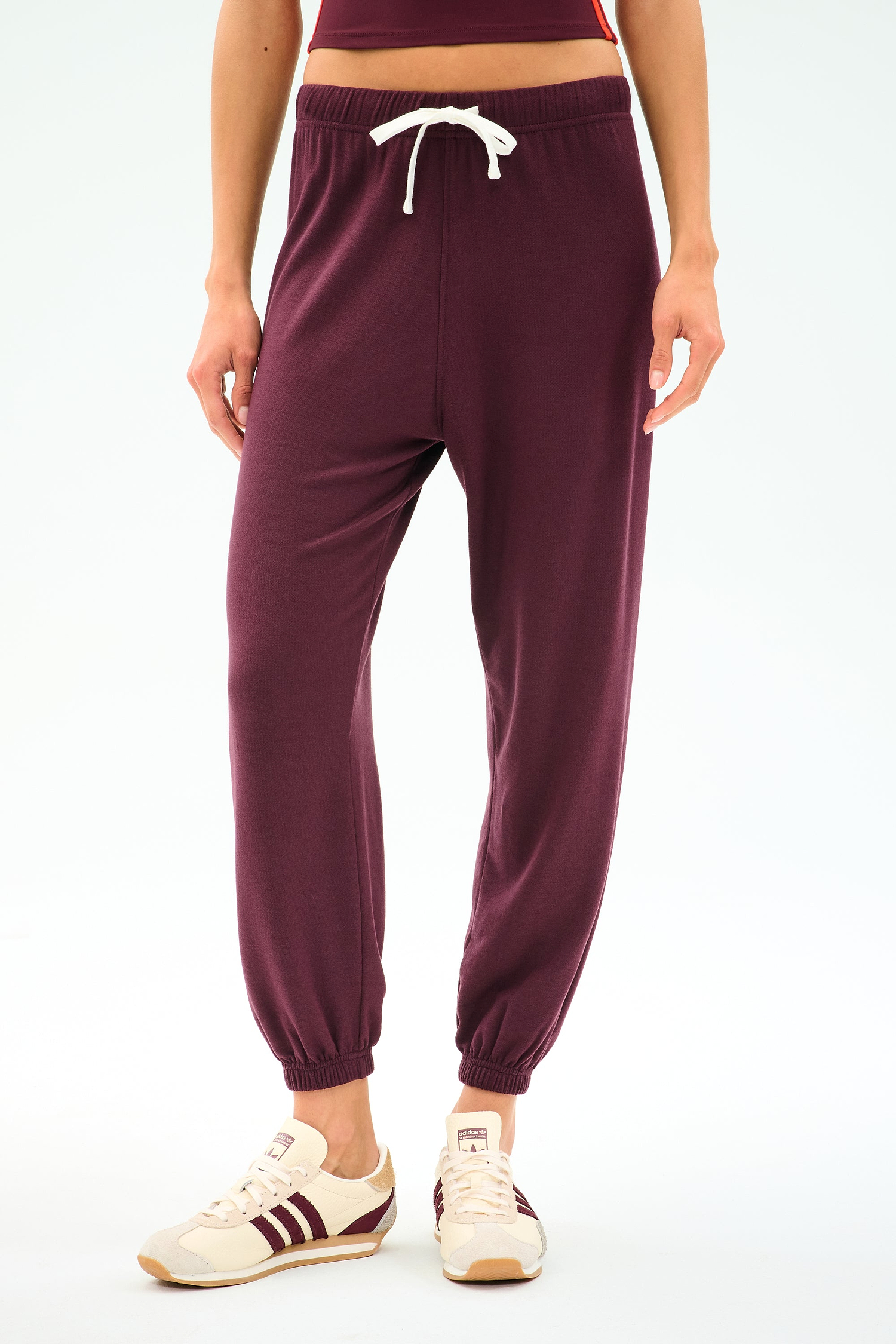 A person is wearing SPLITS59's Andie Oversized Fleece Sweatpant in Dk Cherry, styled with beige sneakers featuring maroon stripes, standing against a plain white background.