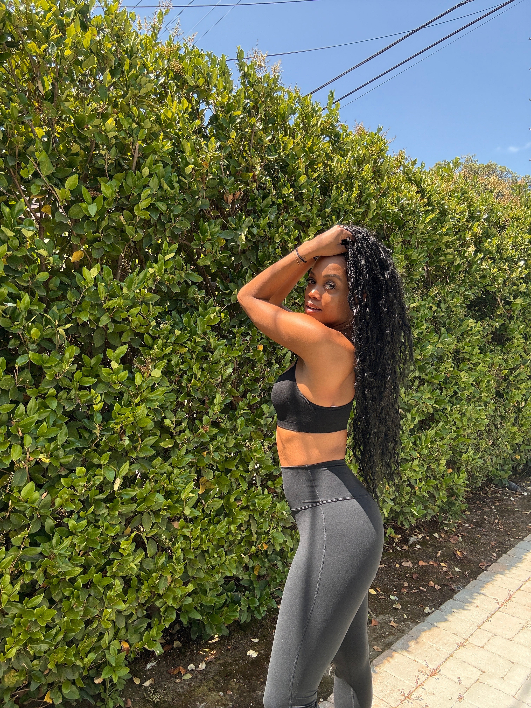 Woman in black sportswear standing outdoors on a paved path next to a tall green hedge, with one hand on her head, looking at the camera.