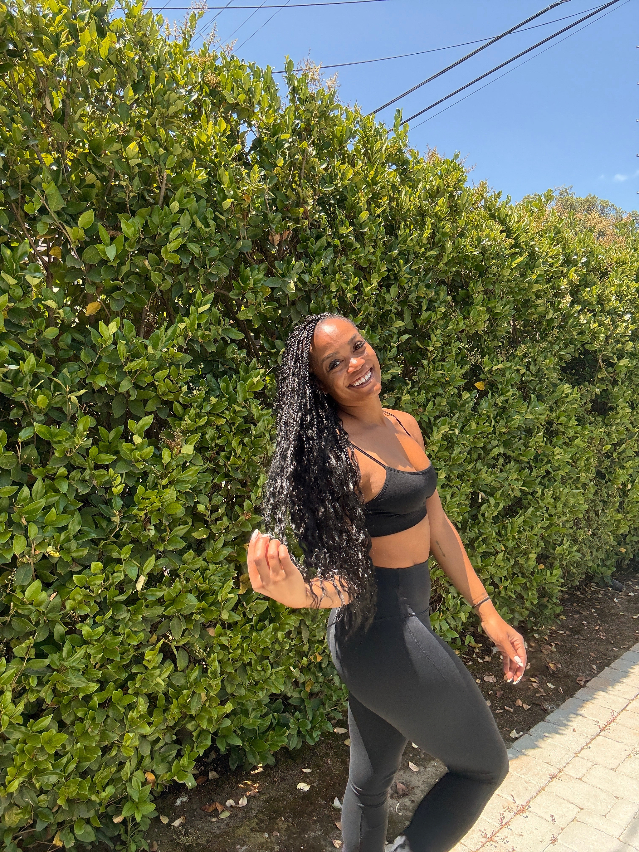 A woman in black athletic wear smiles while standing on a paved path next to a tall green hedge under a sunny sky.