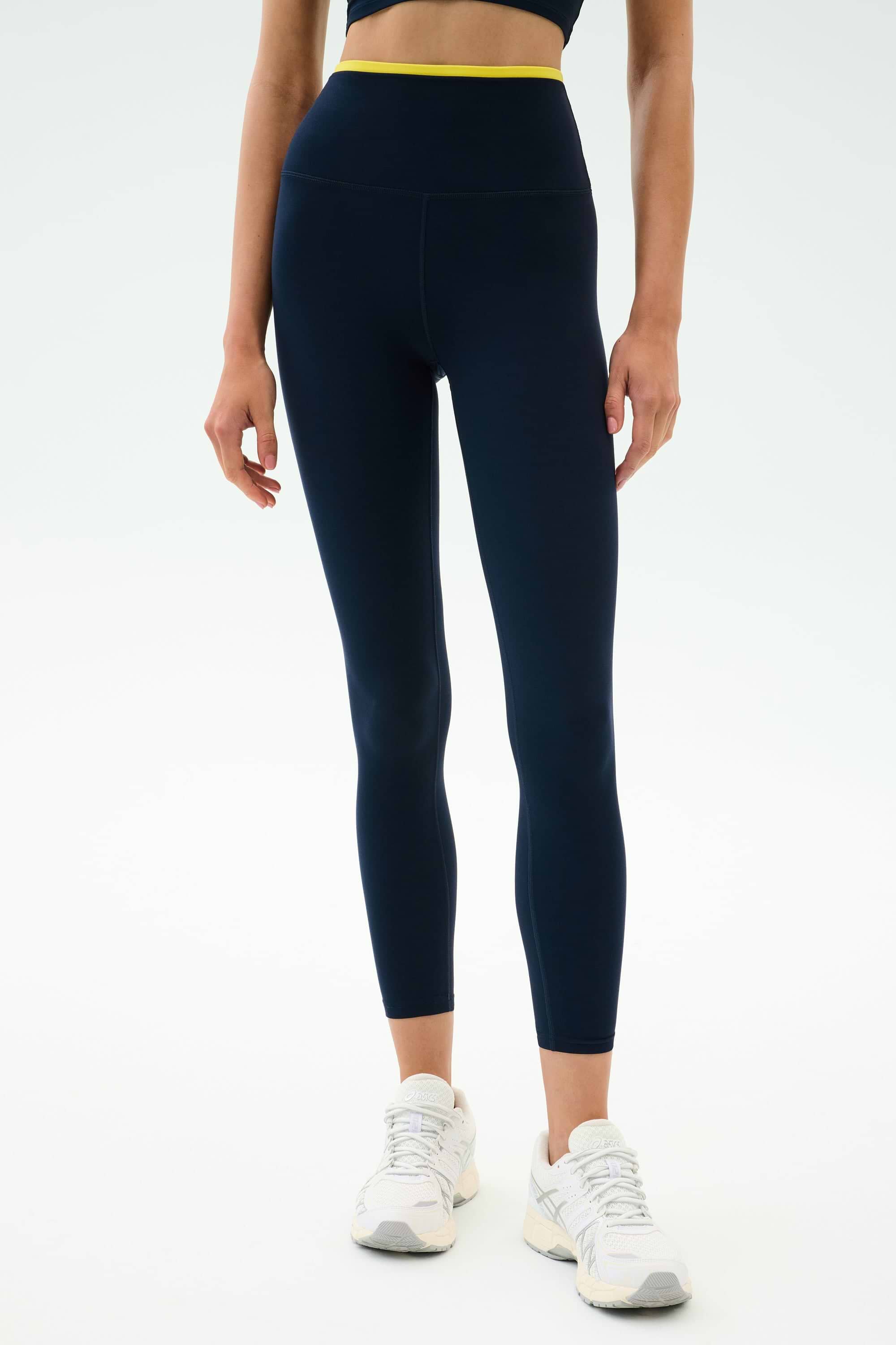 A person stands against a white background in SPLITS59 Dual High Waist Airweight 7/8 leggings in Indigo/Ray—navy with a yellow waistband—paired with white sneakers and a dark crop top, ideal for hot yoga sessions.