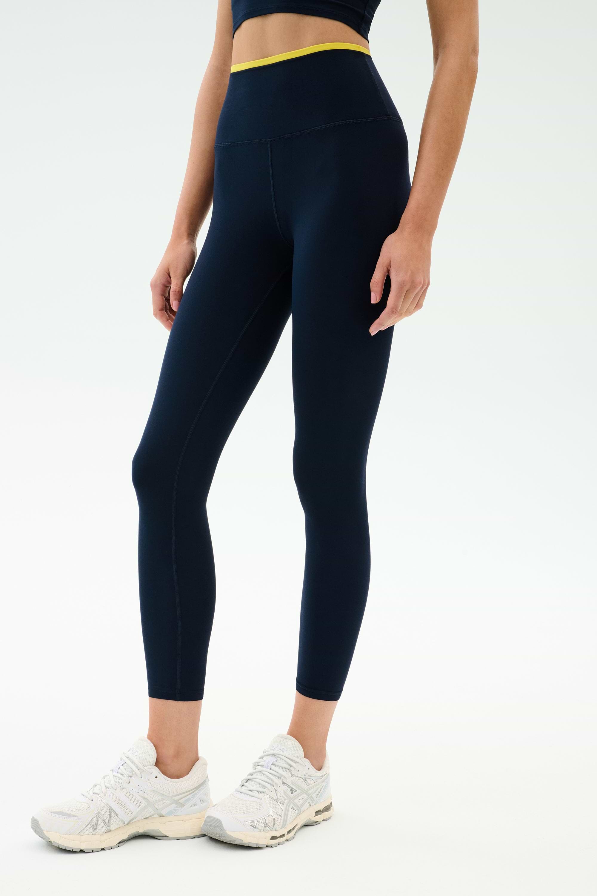 A person stands against a plain background wearing SPLITS59 Dual High Waist Airweight 7/8 leggings in Indigo/Ray with a yellow waistband, white athletic shoes, and a matching navy top—ideal for hot yoga.