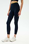 A person stands against a plain background wearing SPLITS59 Dual High Waist Airweight 7/8 leggings in Indigo/Ray with a yellow waistband, white athletic shoes, and a matching navy top—ideal for hot yoga.