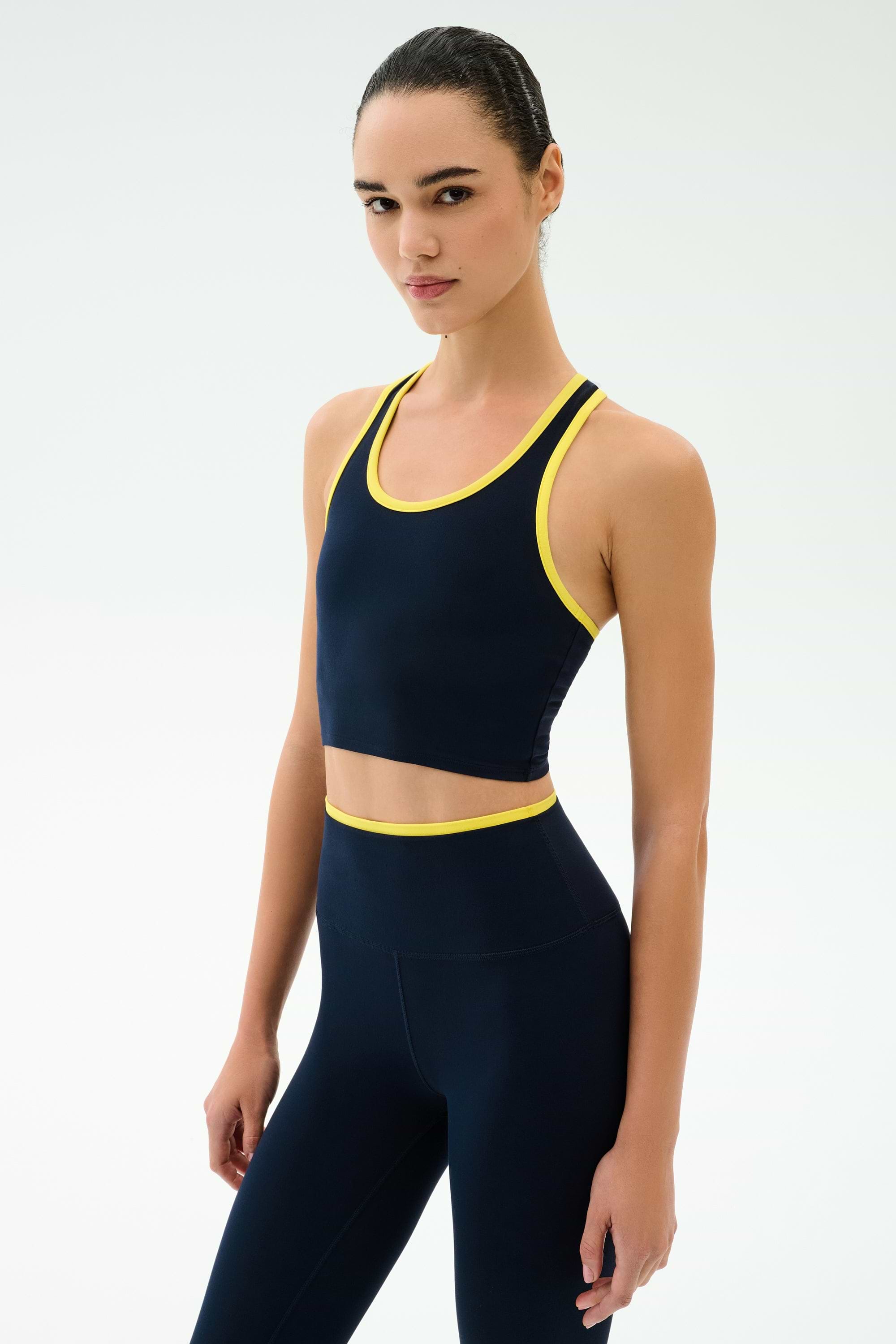 A woman poses against a plain backdrop in the SPLITS59 Amber Airweight Bralette in Indigo/Ray, featuring a scoop neck and yellow trim. Made from Airweight performance fabric, it's ideal for hot yoga sessions.