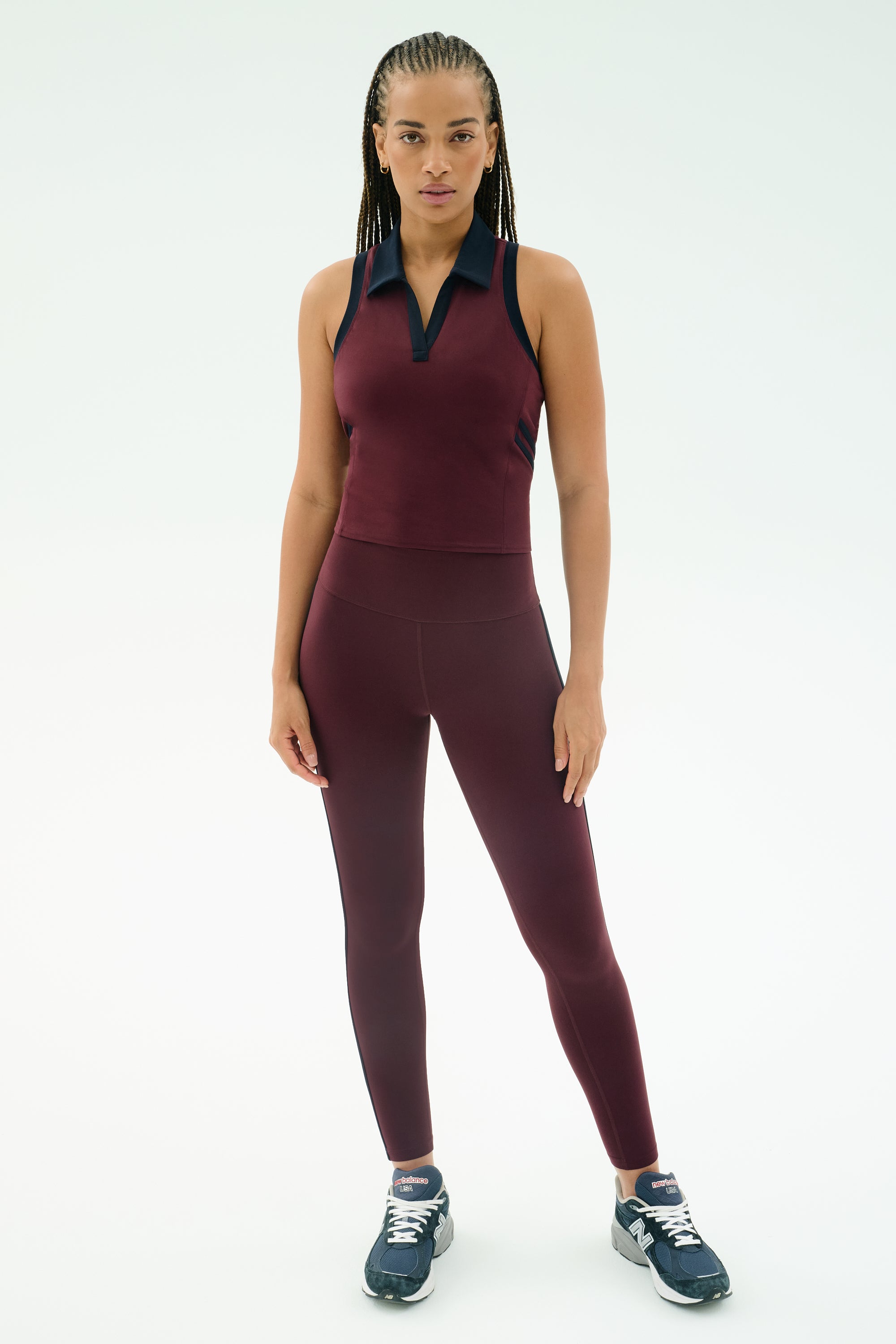 A woman faces forward in SPLITS59's Amber Airweight High Waist 7/8 leggings and top in Dark Cherry/Indigo, crafted from soft nylon spandex, paired with navy sneakers against a white background.