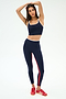 A woman, arms raised overhead, stands on a white background wearing the SPLITS59 Cait Rigor Bralette in Indigo/White with matching leggings featuring red stripes and red sneakers. The set is crafted from stretch fabric.