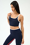 A woman wears the SPLITS59 Cait Rigor Bralette in Indigo/White, styled with matching high-waisted leggings featuring white trim and a red stripe, standing against a plain light background.