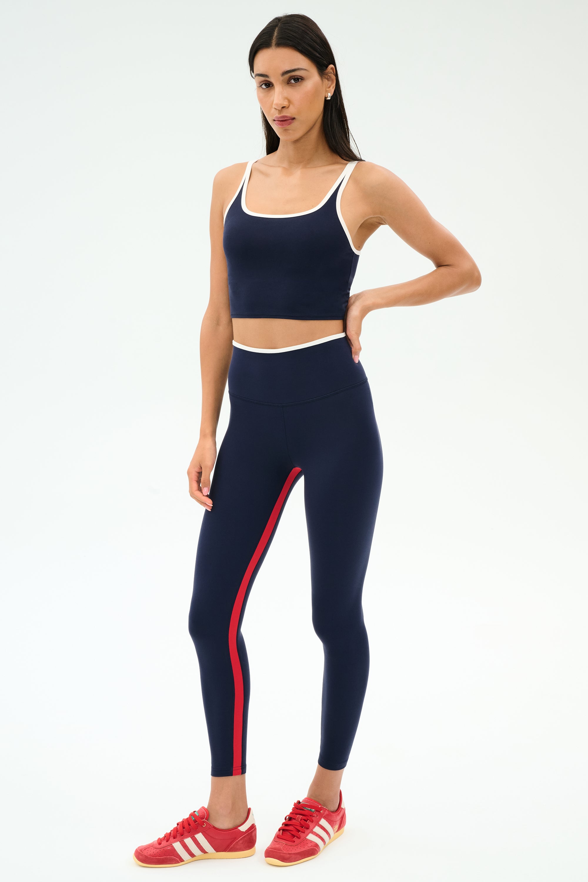 A woman wears the SPLITS59 Cait Rigor Bralette in Indigo/White with matching leggings and red Adidas sneakers, standing against a plain white background.