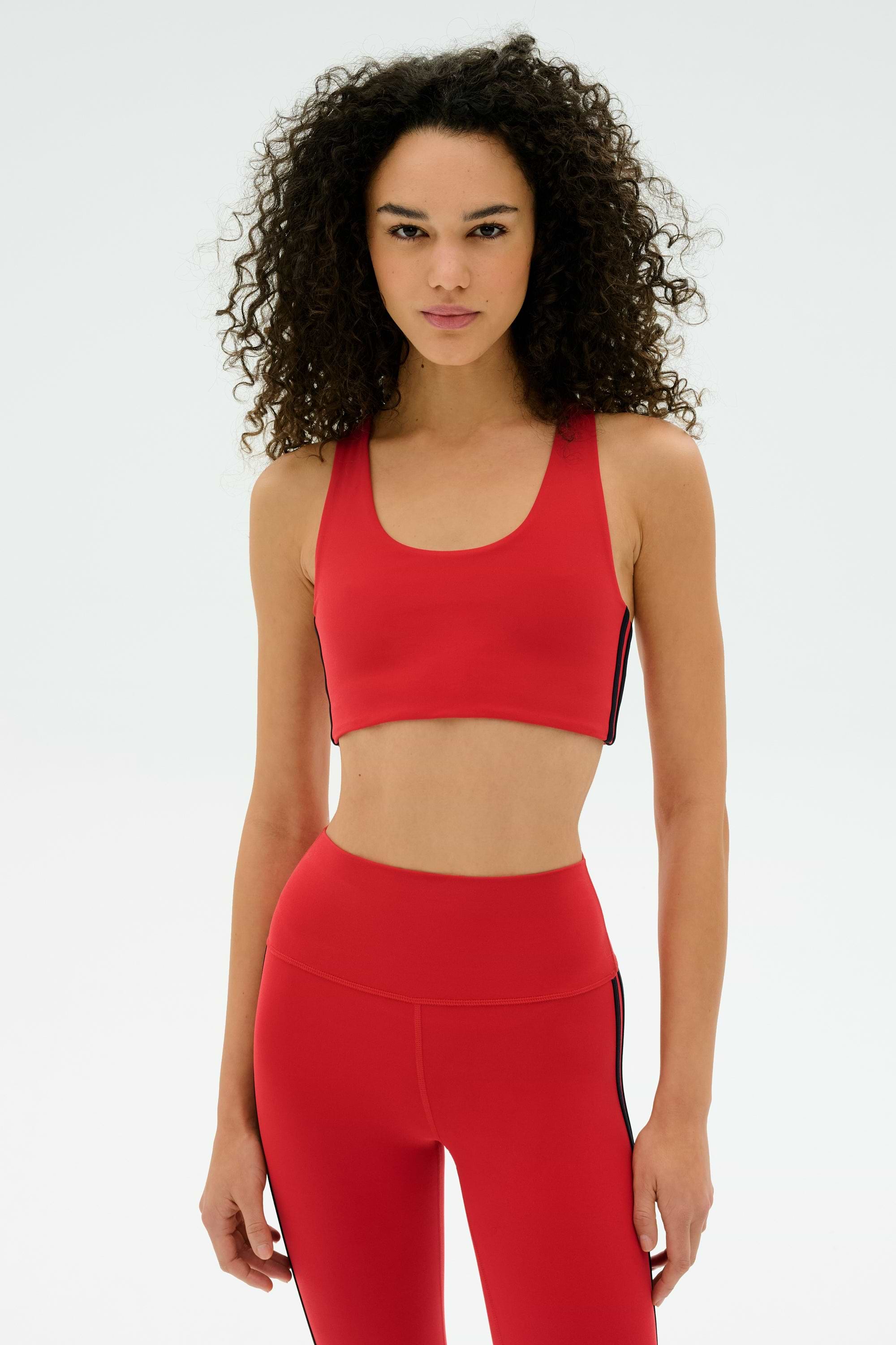 A woman with curly hair wears the SPLITS59 Ella Airweight Bra in Pirate Red/Indigo, paired with high-waisted leggings, both made from stretchy spandex, standing against a plain white background.
