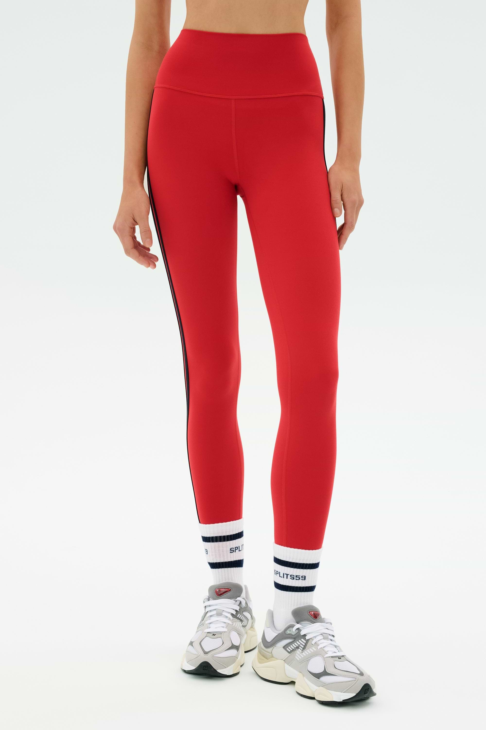 A person wearing SPLITS59 Ella High Waist Airweight 7/8 leggings in Pirate Red/Indigo, white socks with black stripes and text, and white athletic sneakers, standing against a plain background.
