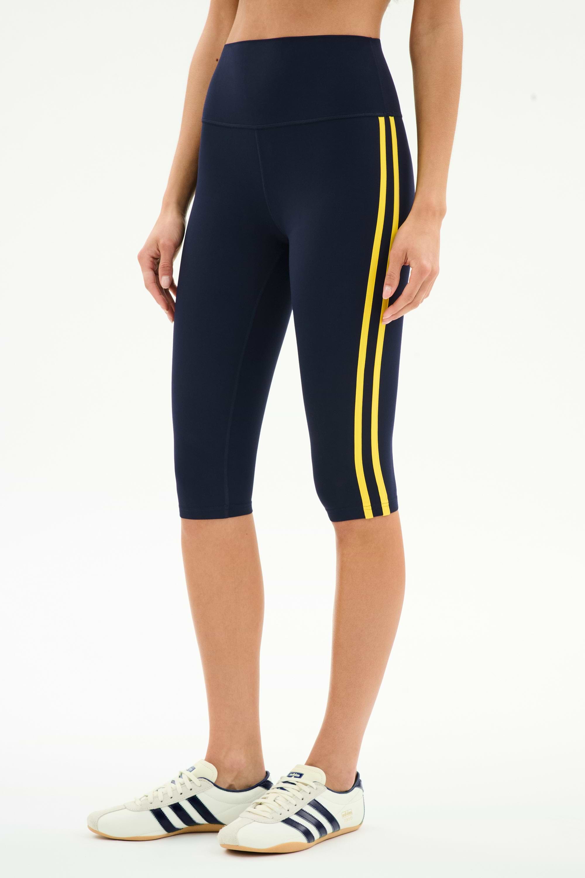 Someone sports SPLITS59’s Ella High Waist Airweight Capri in Indigo/Bright Yellow with white Adidas sneakers featuring navy stripes—a stylish, comfortable outfit perfect for hot yoga or active outings.