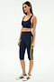 A woman wears the SPLITS59 Ella High Waist Airweight Capri in Indigo/Bright Yellow with a navy sports bra and white sneakers with blue stripes, standing against a plain background—ideal for hot yoga or active days.