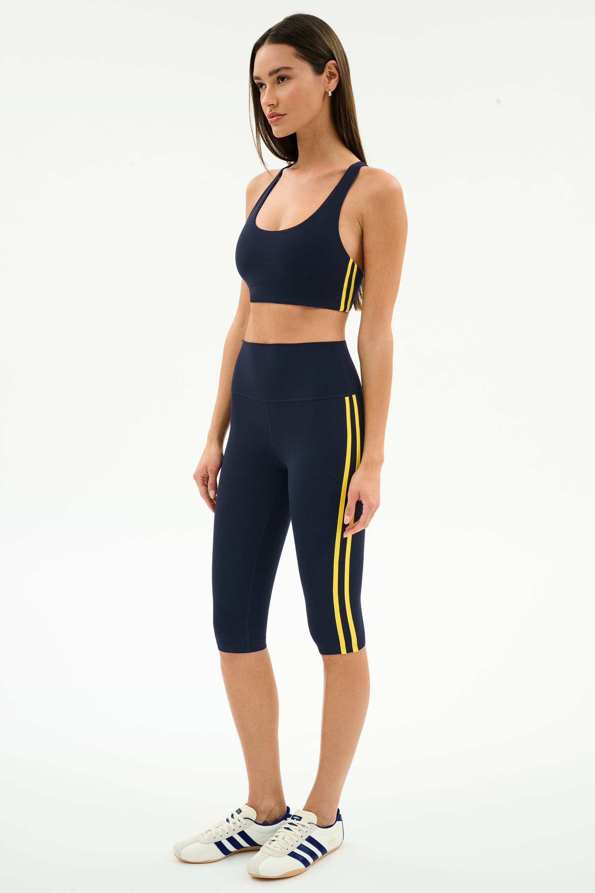 A woman wears the SPLITS59 Ella High Waist Airweight Capri in Indigo/Bright Yellow with a navy sports bra and white sneakers with blue stripes, standing against a plain background—ideal for hot yoga or active days.