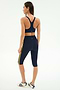 A woman stands facing away in SPLITS59's Ella High Waist Airweight Capri in Indigo/Bright Yellow with a matching sports bra and white sneakers, perfect for hot yoga, set against a plain white background.