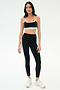 A woman stands against a plain background wearing the SPLITS59 Emerson Airweight Bra in Black/Ice, paired with matching black leggings featuring a white stripe and white athletic shoes—ideal for Pilates or hot yoga.