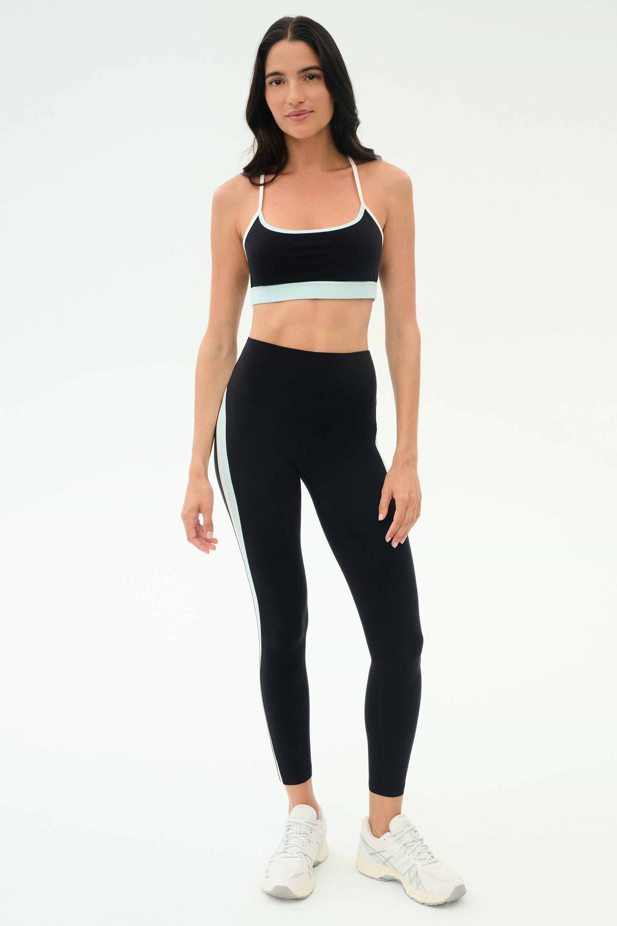 A woman faces forward in a black sports bra, SPLITS59 Emerson Airweight High Waist leggings in Black/Ice with a white side stripe—ideal for hot yoga or Pilates—and white athletic shoes.