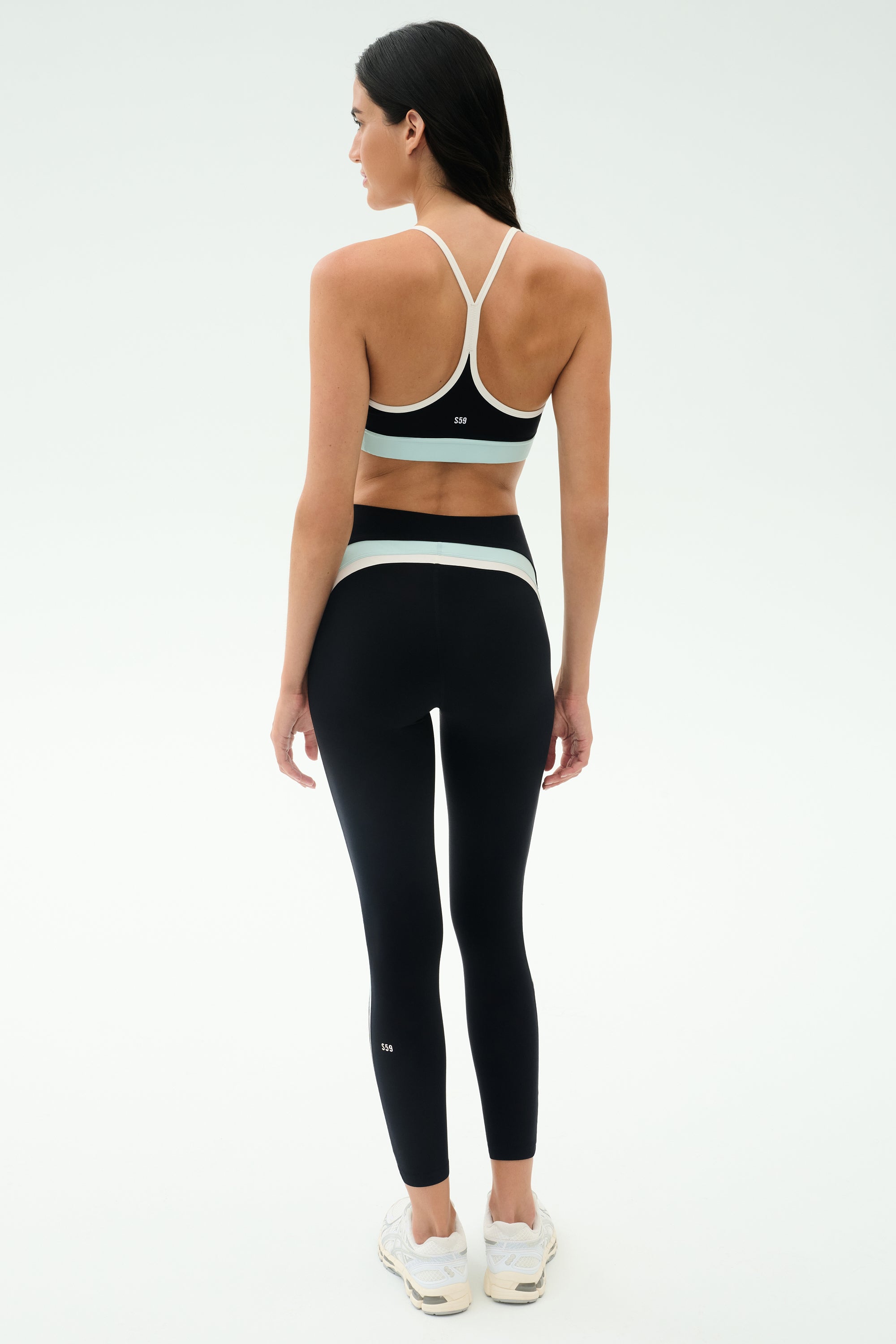 A woman faces away, wearing the SPLITS59 Emerson Airweight Bra in Black/Ice and high-waisted leggings with white accents, plus white sneakers—perfect for Pilates or hot yoga—set against a plain light background.