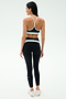 A woman faces away, wearing the SPLITS59 Emerson Airweight Bra in Black/Ice and high-waisted leggings with white accents, plus white sneakers—perfect for Pilates or hot yoga—set against a plain light background.