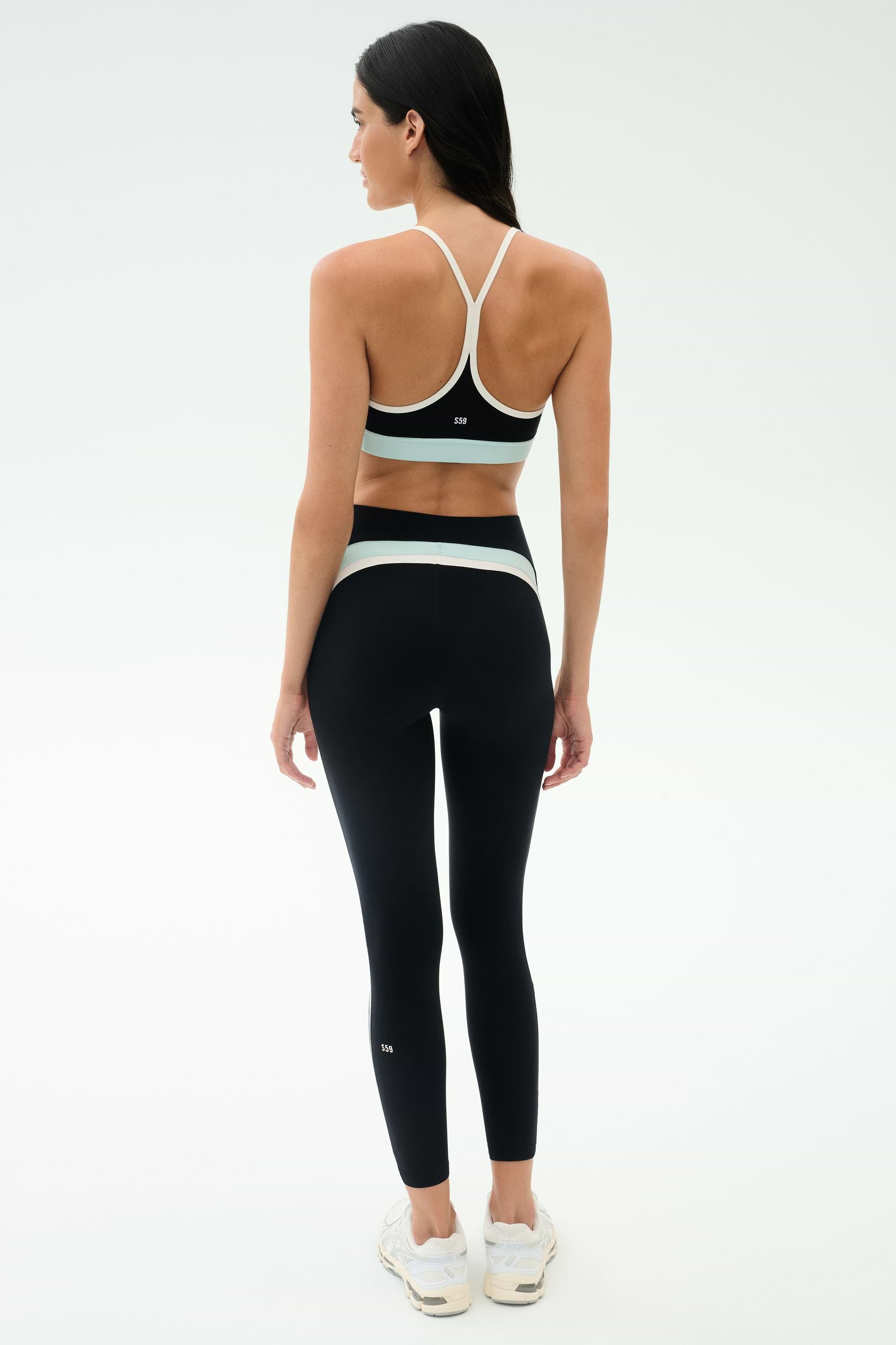A woman faces away in a black SPLITS59 Emerson Airweight High Waist - Black/Ice legging with white accents and a black sports bra, paired with white sneakers—ideal for Pilates or hot yoga—against a plain white background.