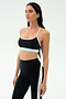 A woman stands in a studio wearing the SPLITS59 Emerson Airweight Bra in Black/Ice, featuring white and light blue trim, paired with high-waisted leggings—an ideal look for Pilates or hot yoga.