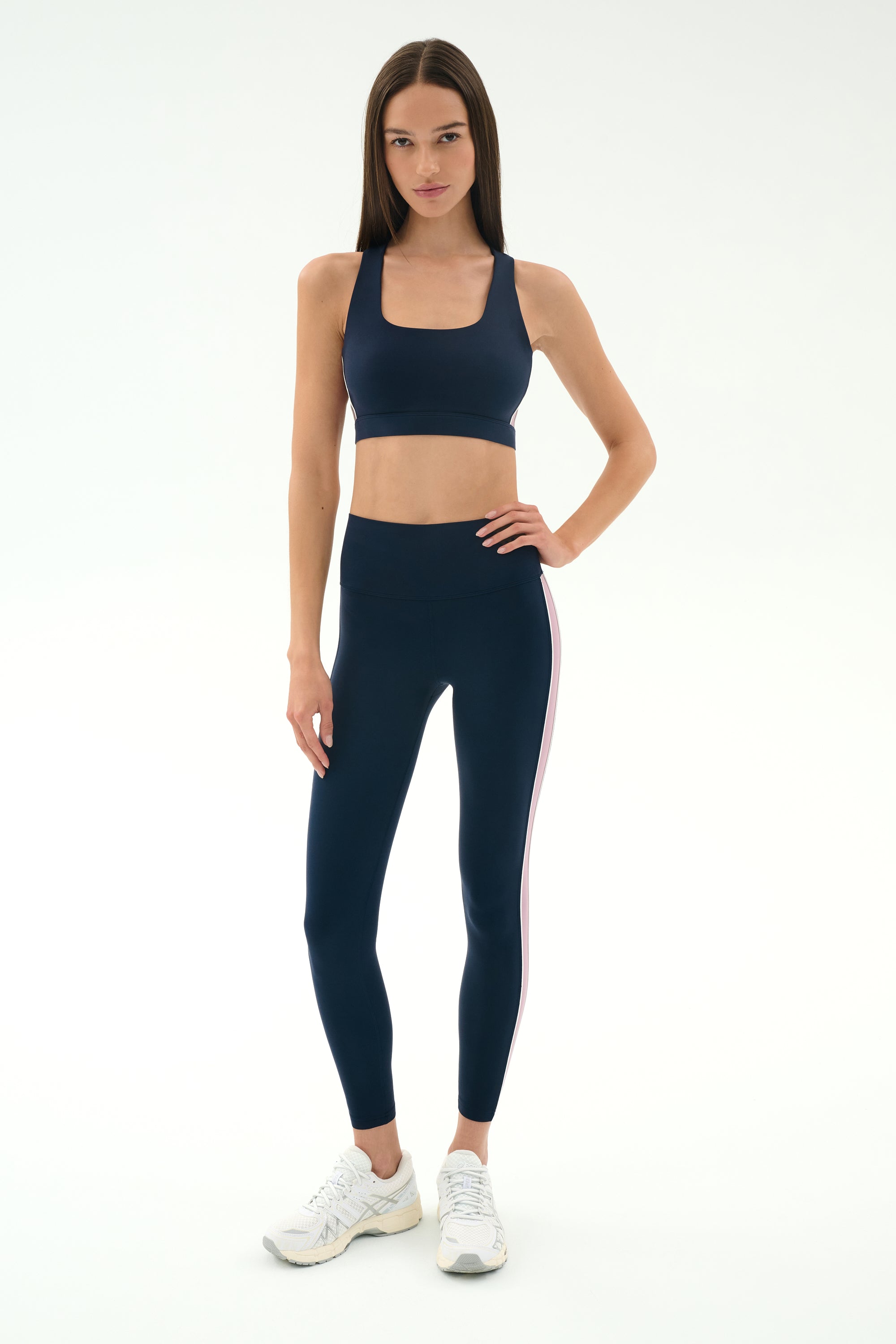 A woman stands against a plain background wearing the SPLITS59 Lexi Rigor Bra in Indigo/Blush, matching leggings with a side stripe, and white sneakers.