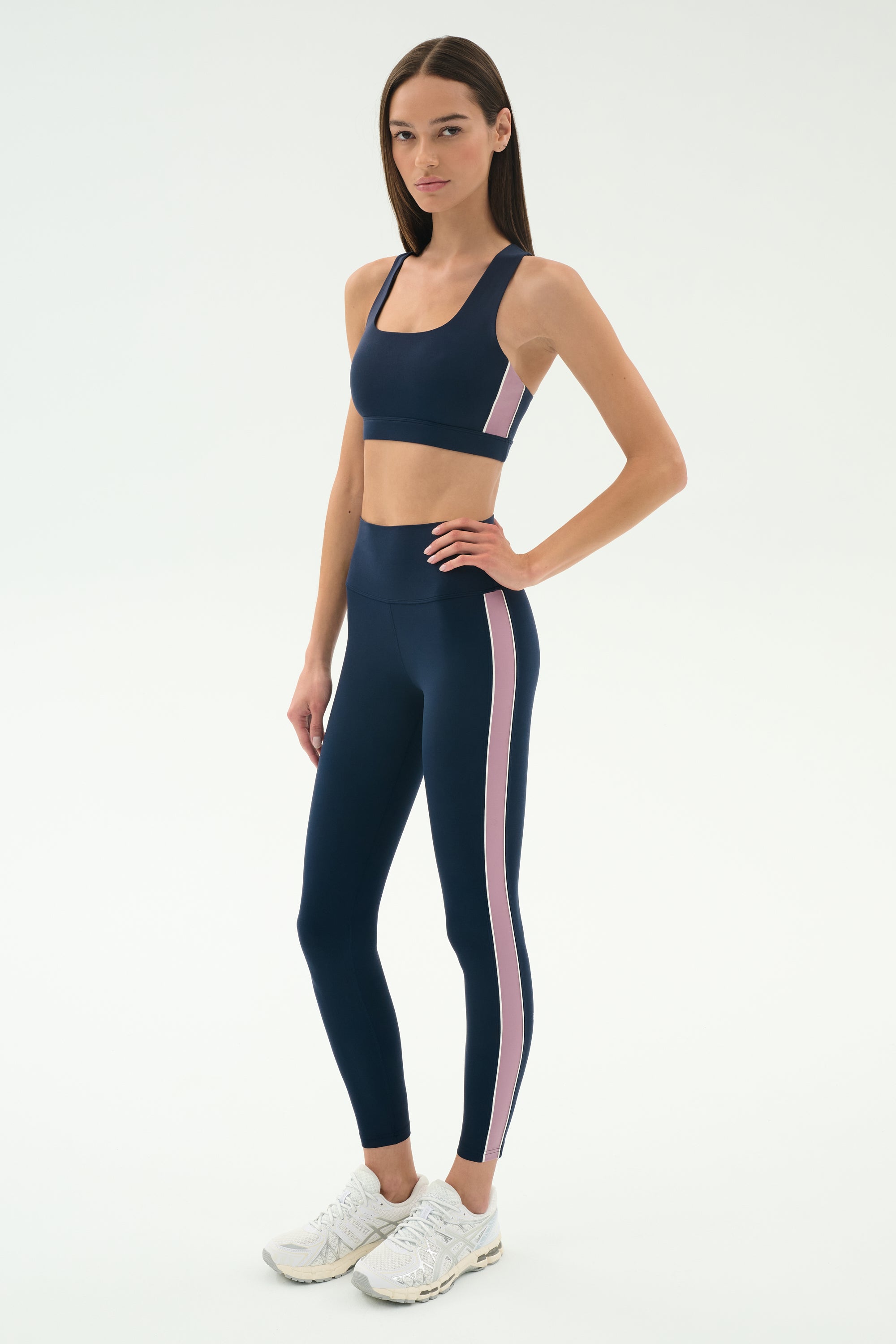 A woman wearing the SPLITS59 Lexi Rigor Bra in Indigo/Blush, featuring a navy blue base with pink side stripes, stands facing the camera against a white background.