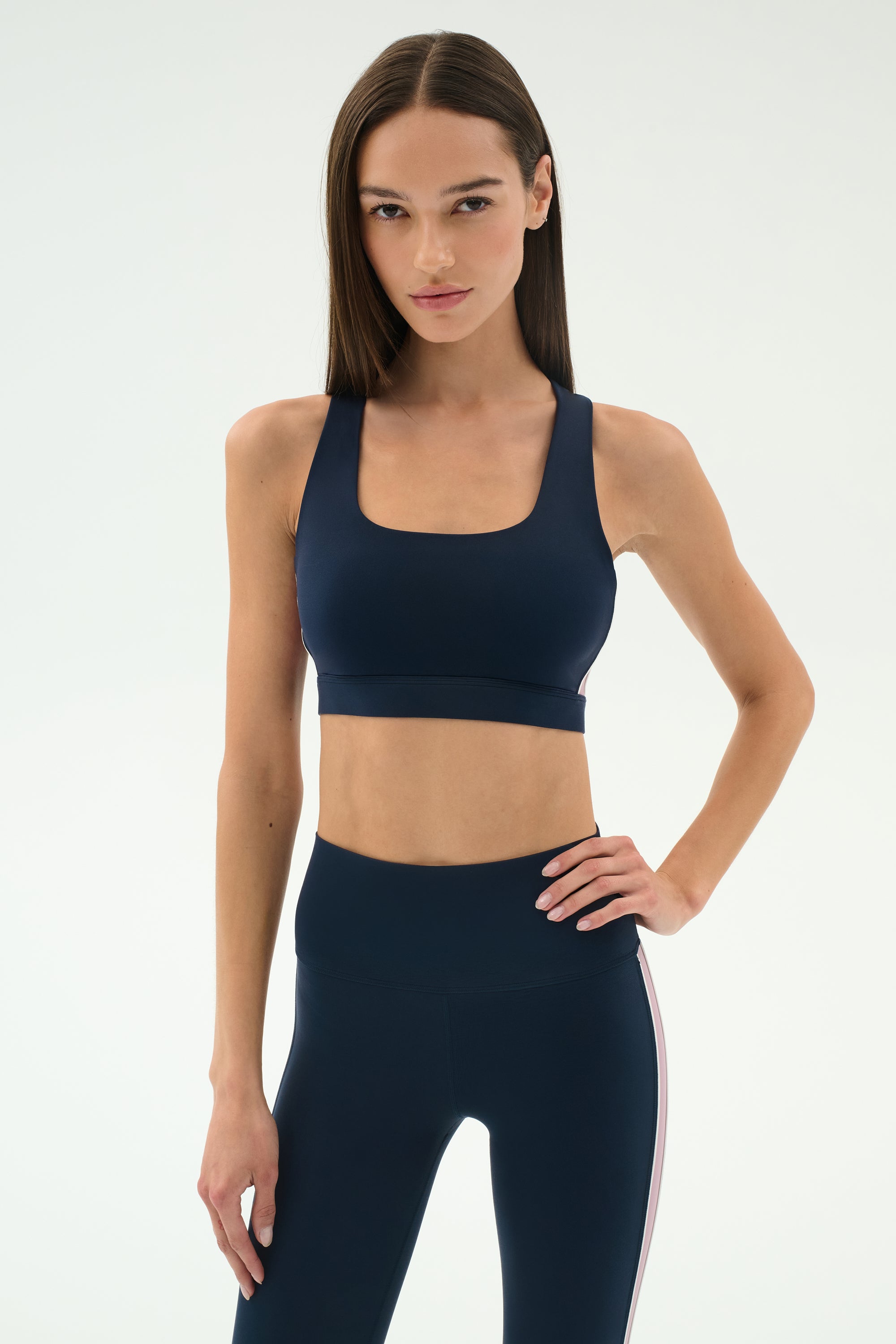 A woman stands against a plain background, wearing the SPLITS59 Lexi Rigor Bra in Indigo/Blush with matching high-waisted leggings, hands on hips.