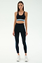 A woman faces forward wearing the SPLITS59 Maci Airweight Bra in Black/Stone Blue with matching high-waisted leggings and white sneakers, set against a plain light background.