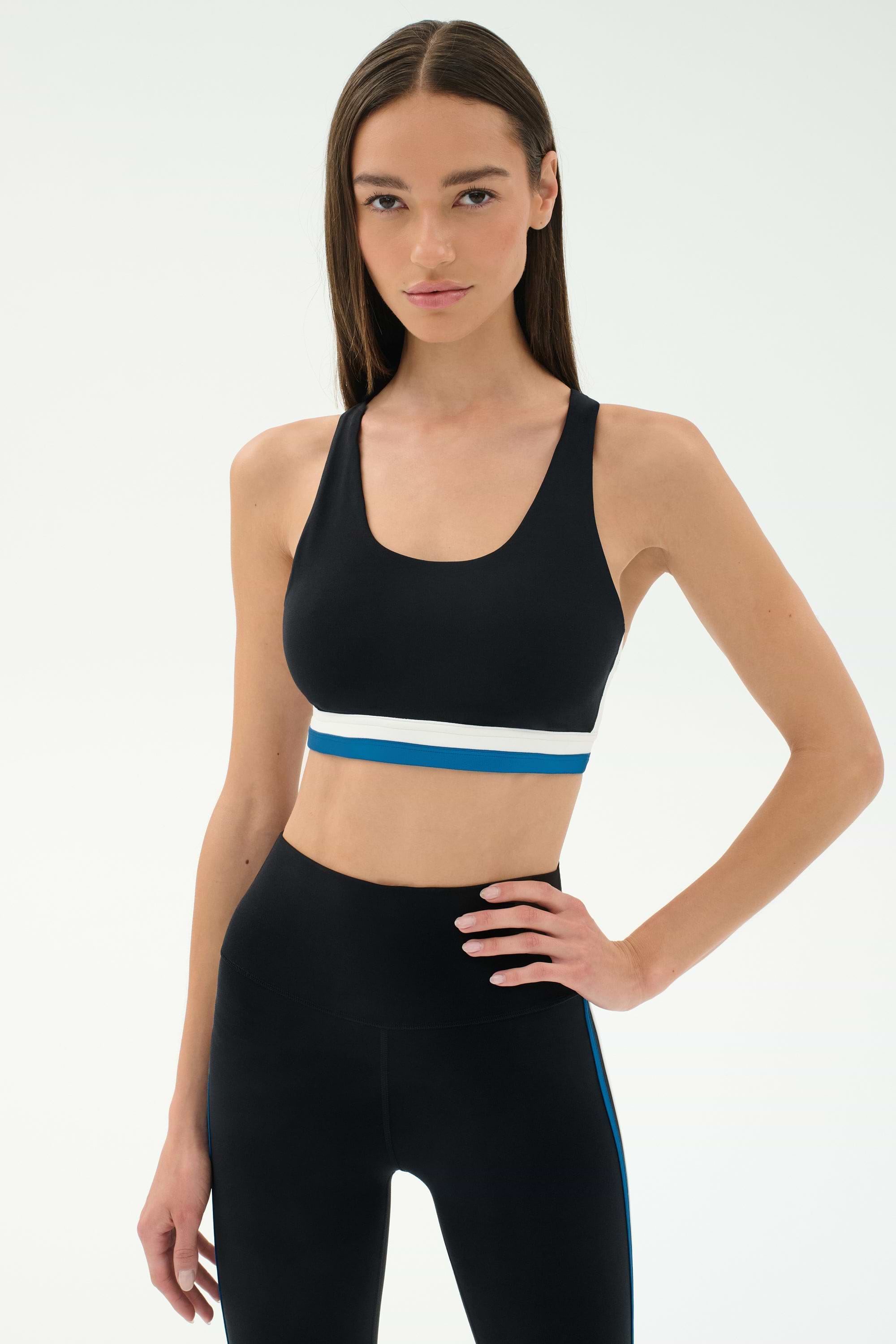 A woman stands with hand on hip wearing the SPLITS59 Maci Airweight Bra in Black/Stone Blue and matching high-waisted leggings, set against a plain light background.
