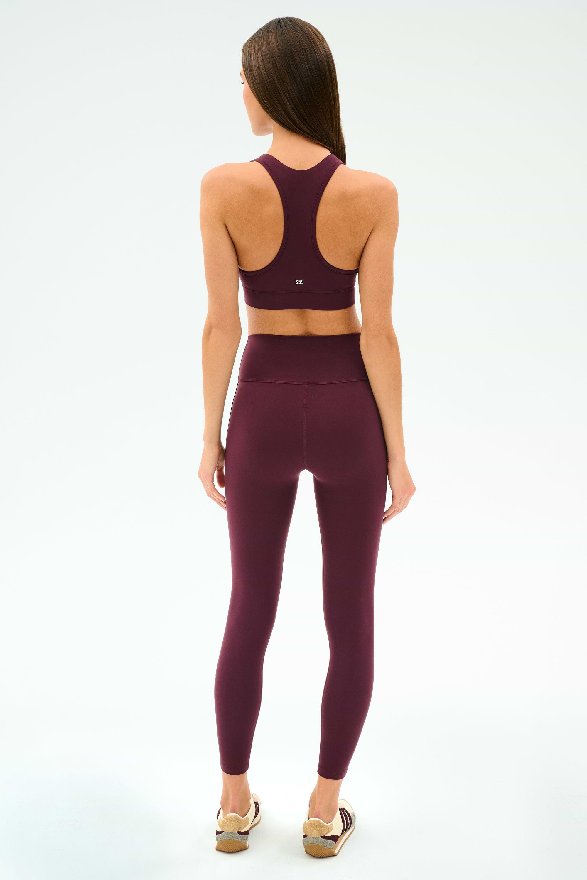 A woman faces away, wearing the SPLITS59 Mia Rigor Bra in Dark Cherry with matching leggings and light sneakers, set against a plain white background.