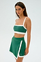 A woman displays her stylish workout outfit, including the SPLITS59 Monah Rigor Bra in Arugula/White and a matching skirt. Posed against a plain background, she stands slightly turned, exuding confidence for her gym workouts.