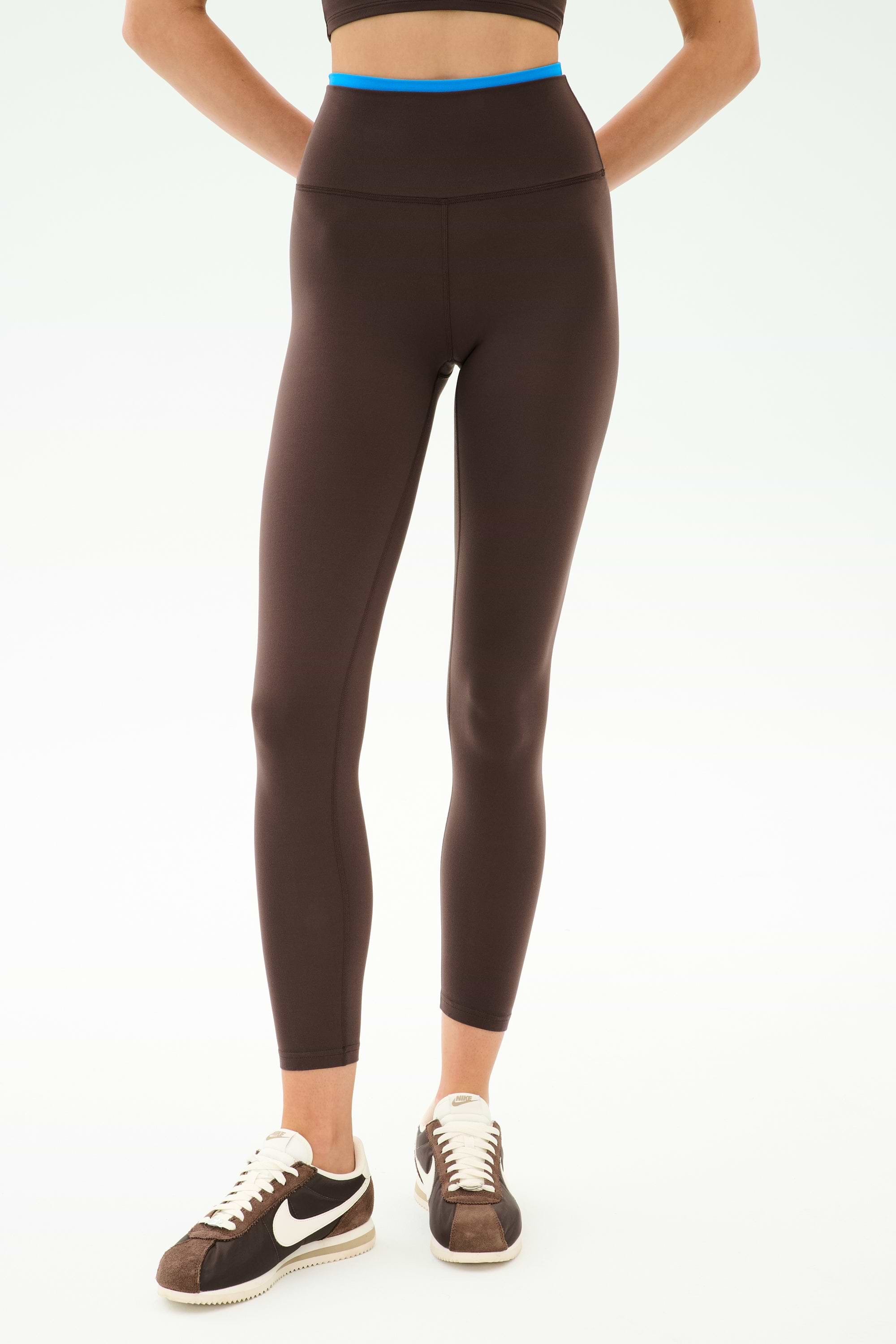 A person wears SPLITS59 Dual High Waist Airweight 7/8 leggings in Dark Chocolate/Classic Blue, paired with a black sports bra and brown and white Nike sneakers, standing against a plain light background—ideal for hot yoga.