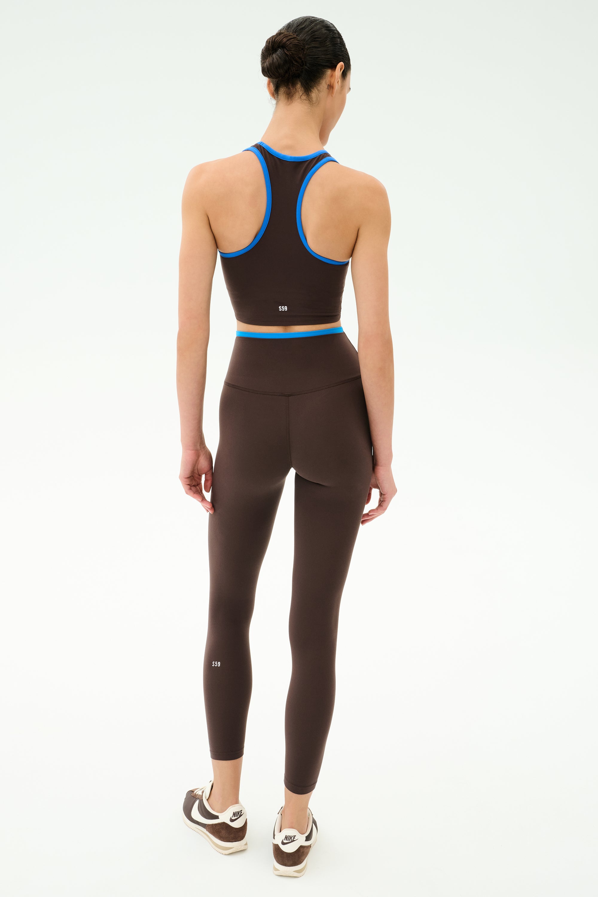 A person stands facing away in SPLITS59 Dual High Waist Airweight 7/8 leggings in Dark Chocolate/Classic Blue and a brown athletic crop top, paired with brown and white sneakers—ideal for hot yoga sessions against a plain light background.
