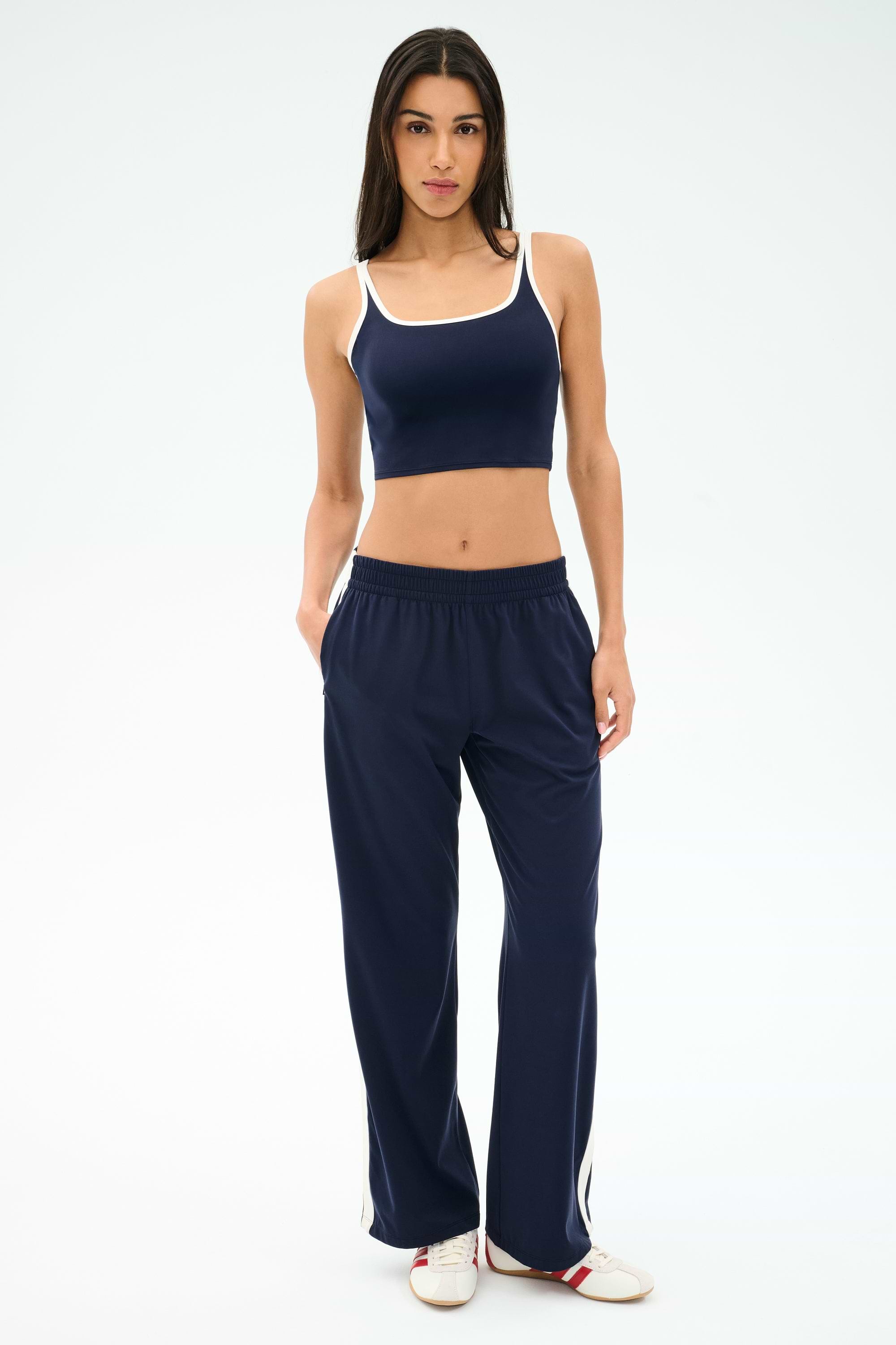 A woman faces forward wearing SPLITS59's Maxie Rigor Track Pant in Indigo/White with white trim, a coordinating navy crop top, and white sneakers.