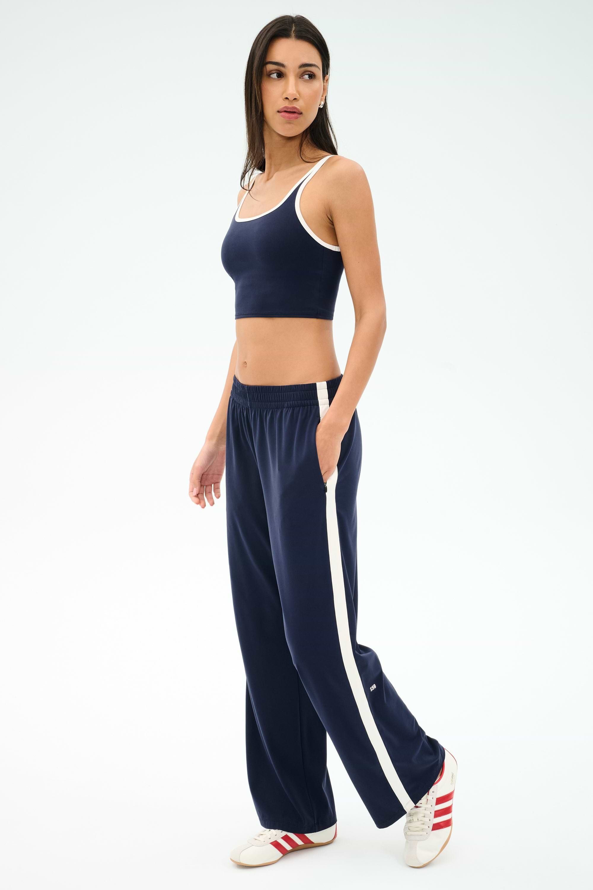 A woman in a navy crop top and SPLITS59 Maxie Rigor Track Pant - Indigo/White, wide-leg pants with white stripes, paired with red and white sneakers, stands against a plain background.