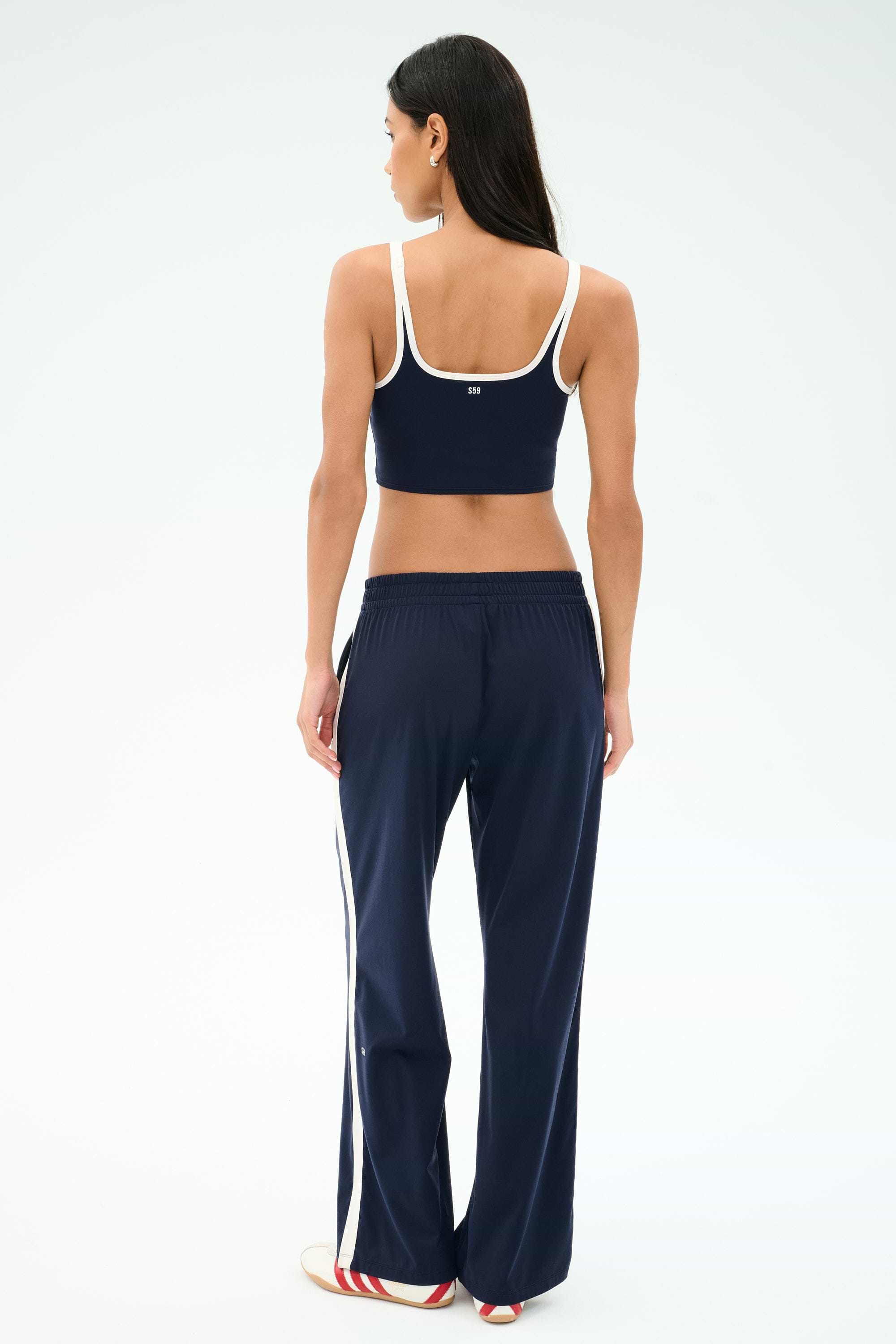 A woman, facing away, wears a navy blue crop top and SPLITS59 Maxie Rigor Track Pant in Indigo/White with white trim and side stripes, against a plain white background.