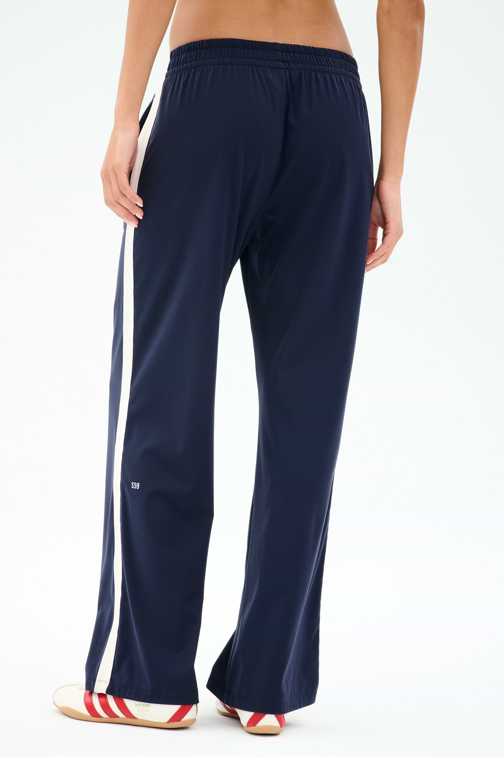 Person wearing SPLITS59 Maxie Rigor Track Pant - Indigo/White with white side stripes and sneakers, shown from the back against a plain light background. Wide leg pants crafted from durable Rigor fabric for all-day comfort.