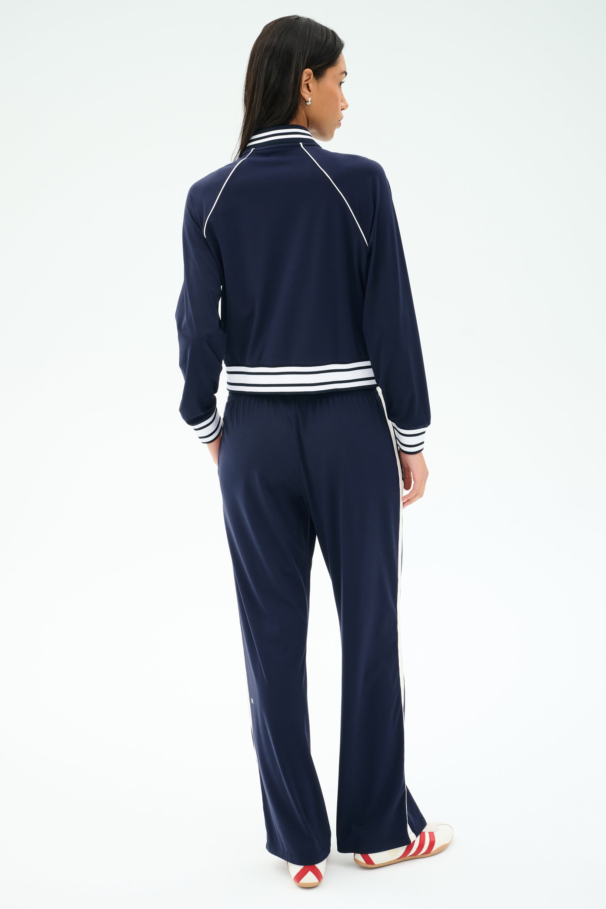 A woman faces away, wearing the SPLITS59 Hayes Varsity Rigor Jacket in indigo, paired with red-and-white sneakers; the stretch fabric tracksuit features white stripes and stands out against a plain light background.