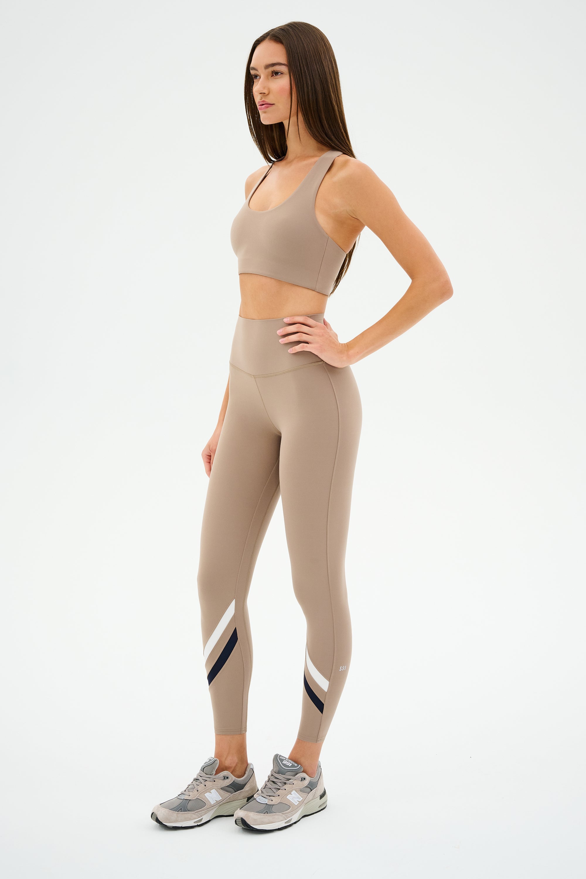 A woman models SPLITS59's Chevron Airweight High Waist 7/8 leggings in Latte/Indigo, highlighting the ultra-luxe fabric with trendy sneakers against a plain backdrop.
