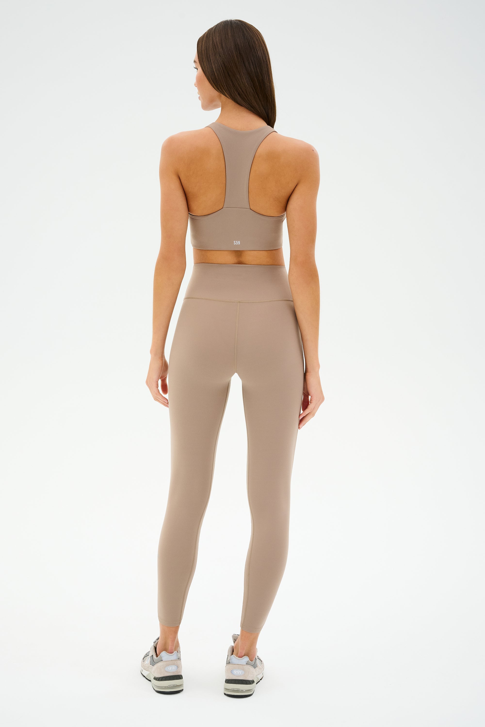 A person in taupe athletic wear and sneakers, showcasing SPLITS59 Chevron Airweight High Waist 7/8 leggings in Latte/Indigo, stands with their back to the camera against a plain white background.