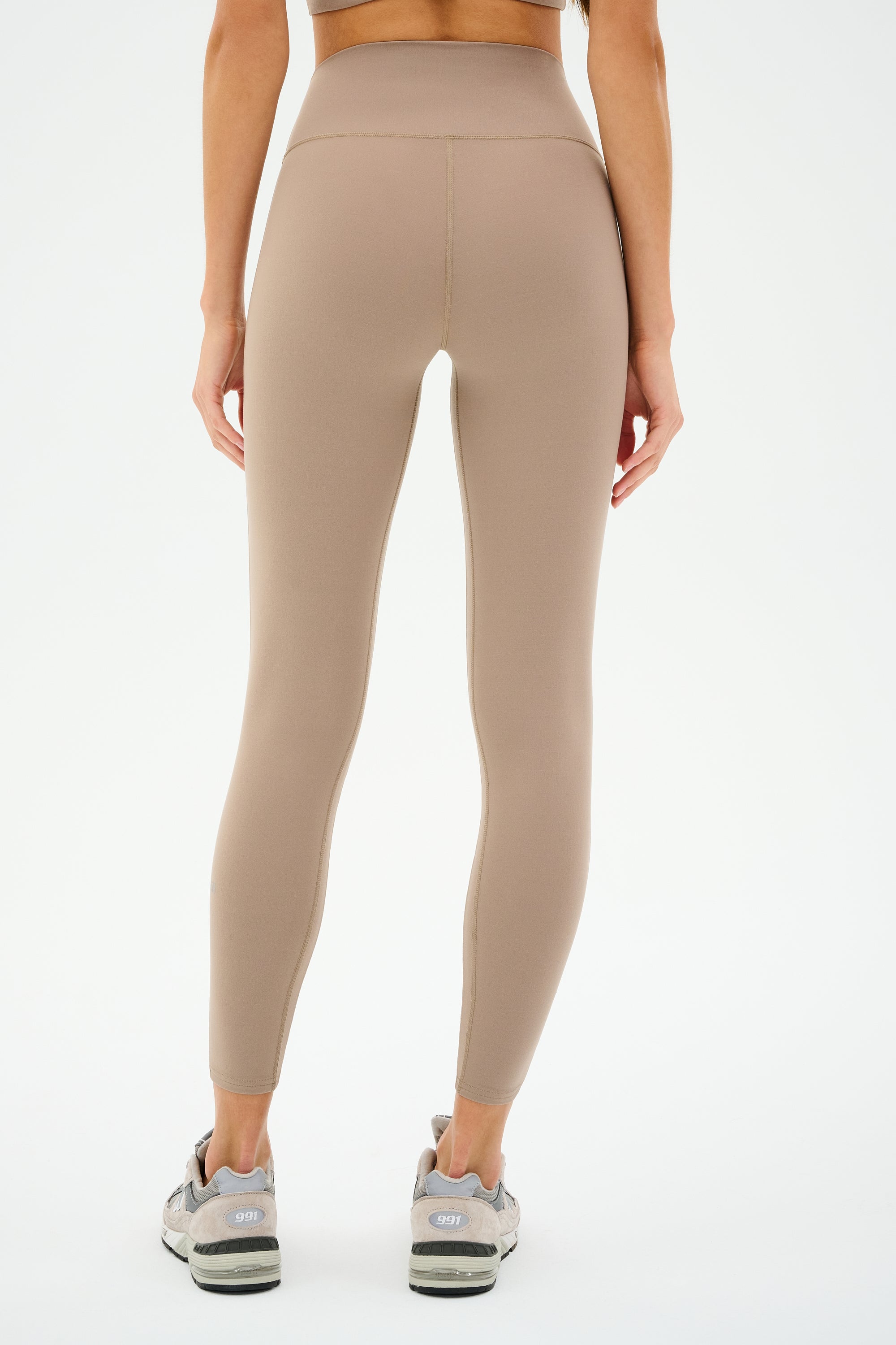 A person stands with their back to the camera against a white backdrop, wearing SPLITS59 Chevron Airweight High Waist 7/8 leggings in Latte/Indigo and gray athletic shoes.